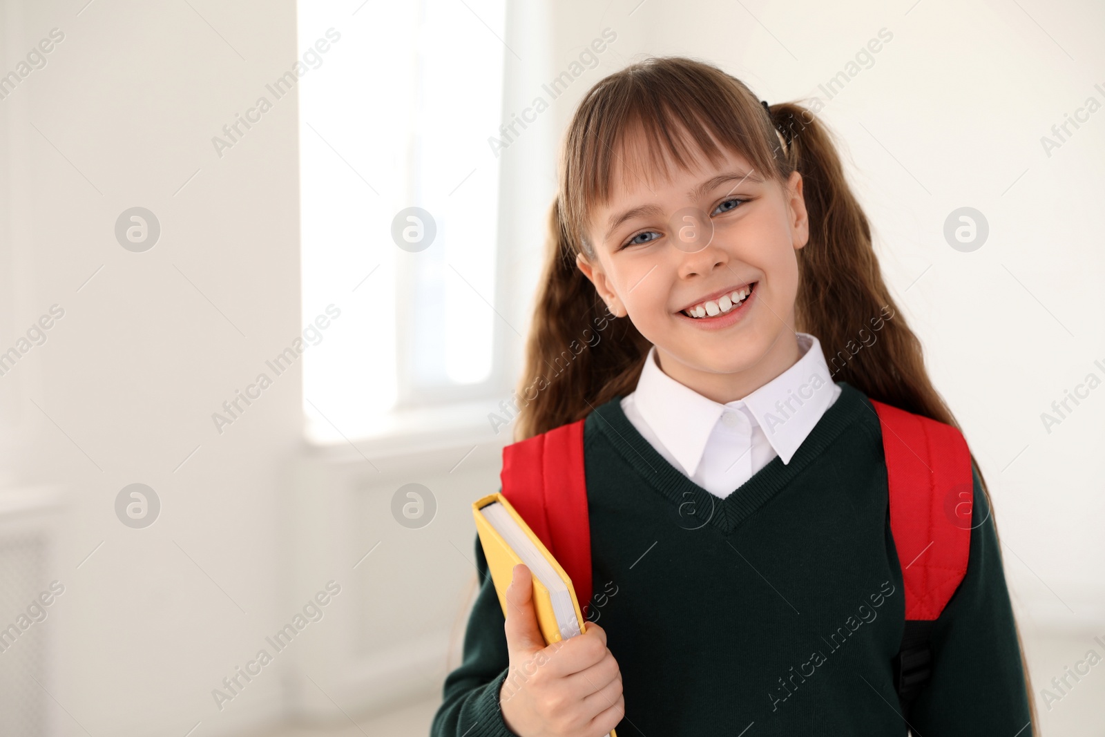 Photo of Portrait of cute girl in school uniform with backpack and book indoors. Space for text