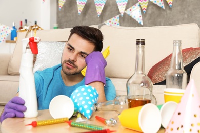 Tired young man with bottle of detergent and rag in messy room after party