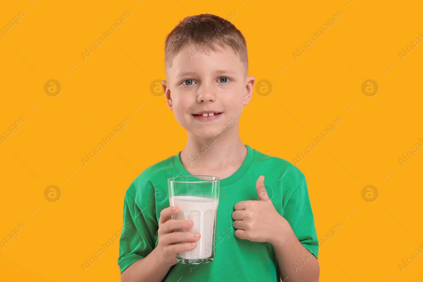 Photo of Cute boy with glass of fresh milk showing thumb up on orange background