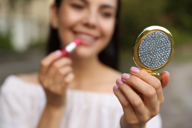 Photo of Woman with cosmetic pocket mirror applying lipstick outdoors, closeup
