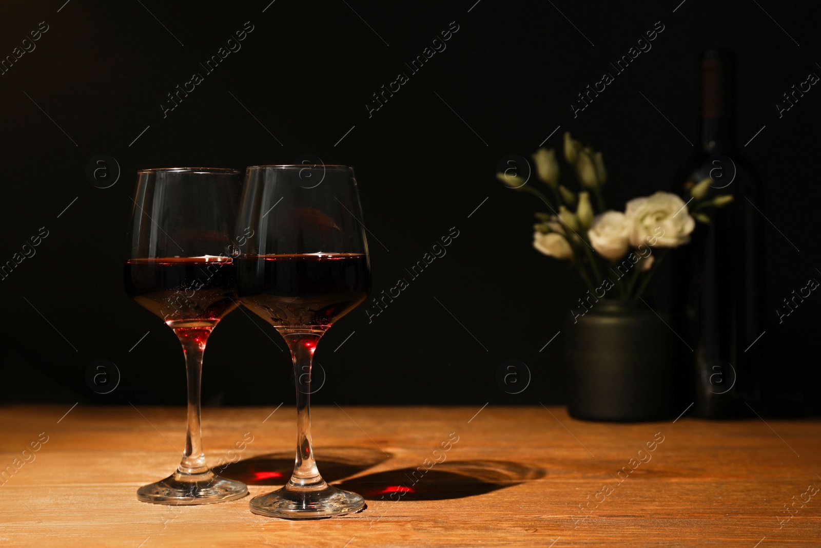 Photo of Glasses of wine and flowers on wooden table near dark wall