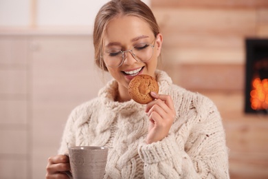 Photo of Beautiful young woman with cup of hot drink and cookie at home. Winter atmosphere
