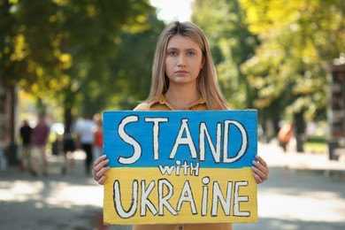 Photo of Sad woman holding poster in colors of national flag and words Stand with Ukraine outdoors