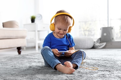 Photo of Cute child with headphones and mobile phone on floor indoors