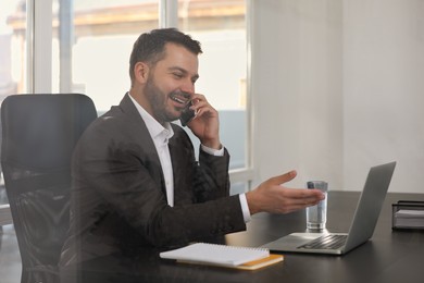 Photo of Happy man using modern laptop while talking on smartphone at black desk in office