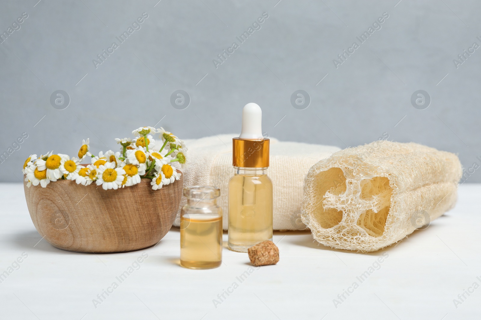 Photo of Essential oil, chamomiles and loofah on white table against grey background