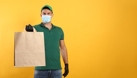 Photo of Courier in medical mask holding paper bag with takeaway food on yellow background, space for text. Delivery service during quarantine due to Covid-19 outbreak