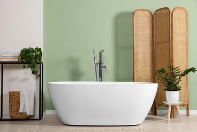 Photo of Modern ceramic tub and beautiful plants near light green wall in bathroom. Interior design