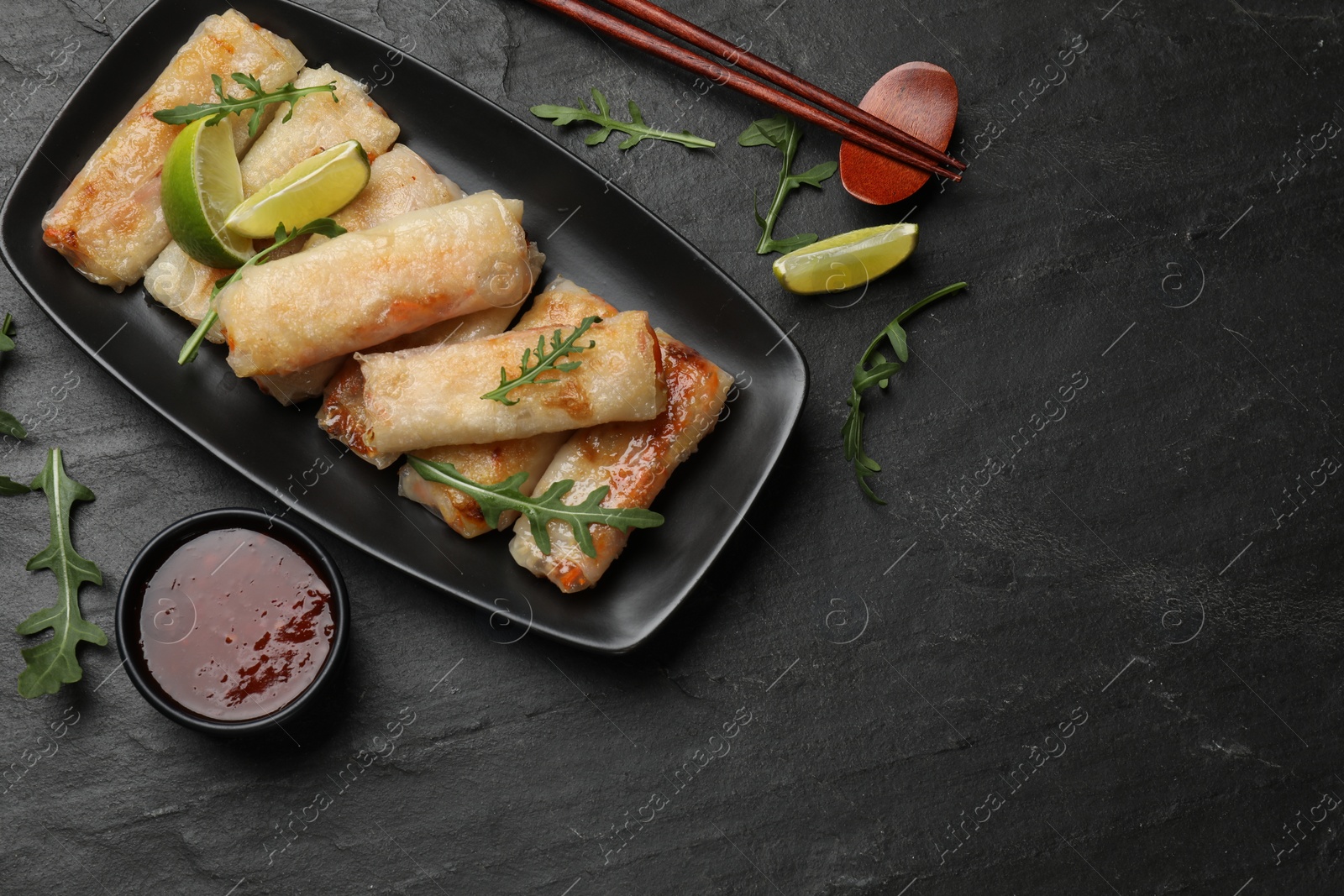 Photo of Tasty fried spring rolls, arugula, lime and sauce on dark textured table, flat lay. Space for text