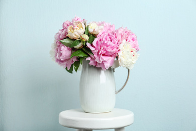 Photo of Beautiful peonies in jug on white stool against light blue background