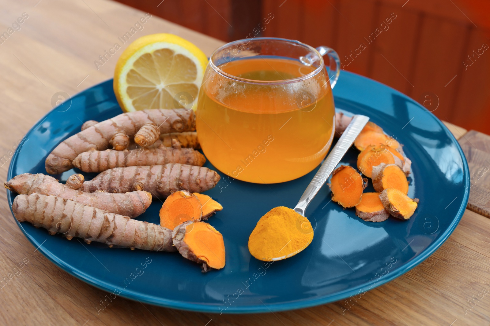 Photo of Plate with glass cup of hot tea, lemon, turmeric powder and roots on wooden table