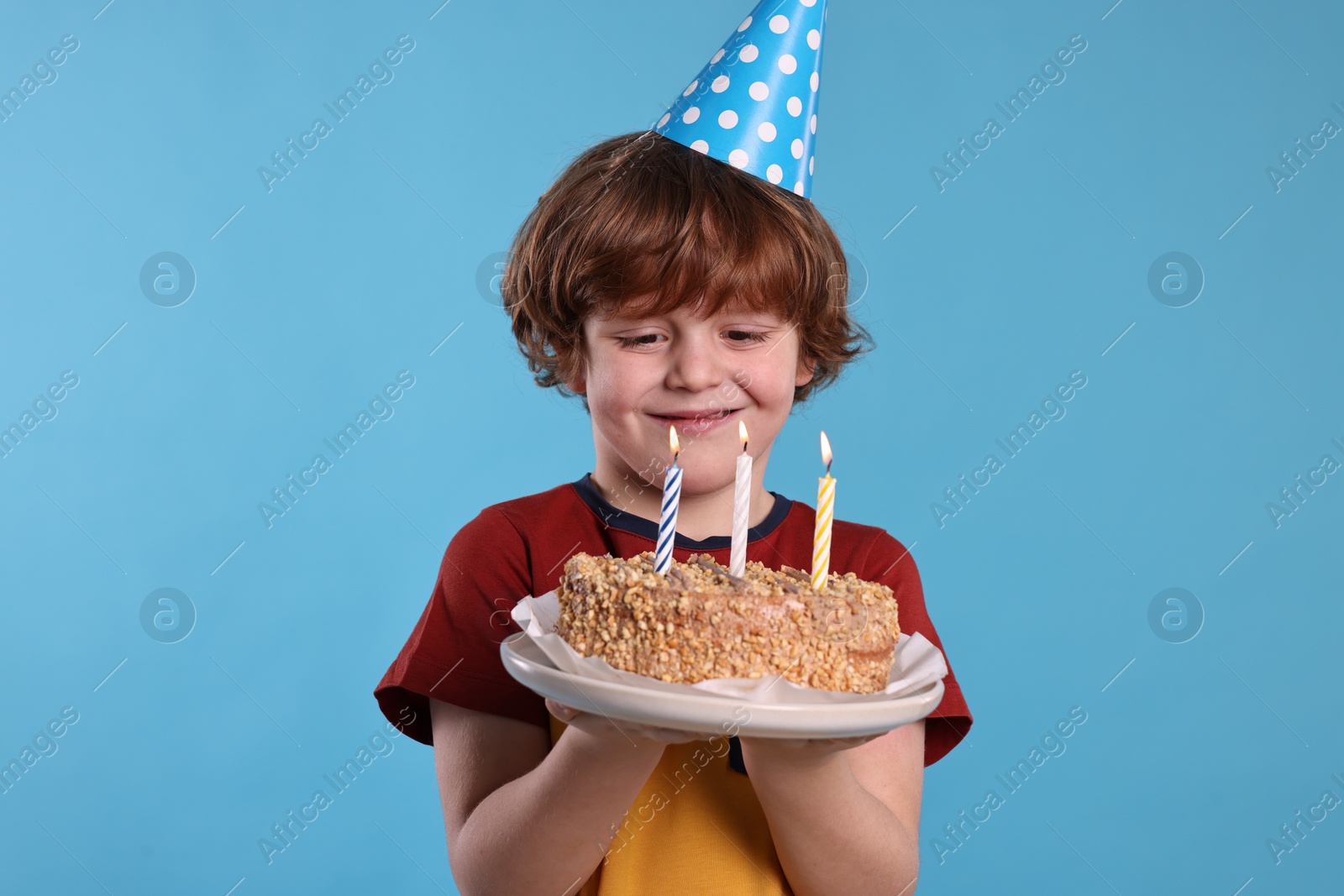 Photo of Birthday celebration. Cute little boy in party hat holding tasty cake with burning candles on light blue background