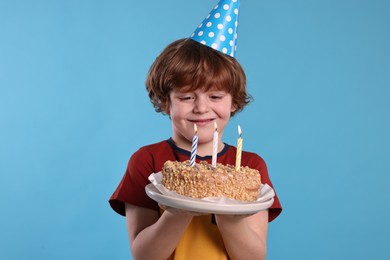 Photo of Birthday celebration. Cute little boy in party hat holding tasty cake with burning candles on light blue background