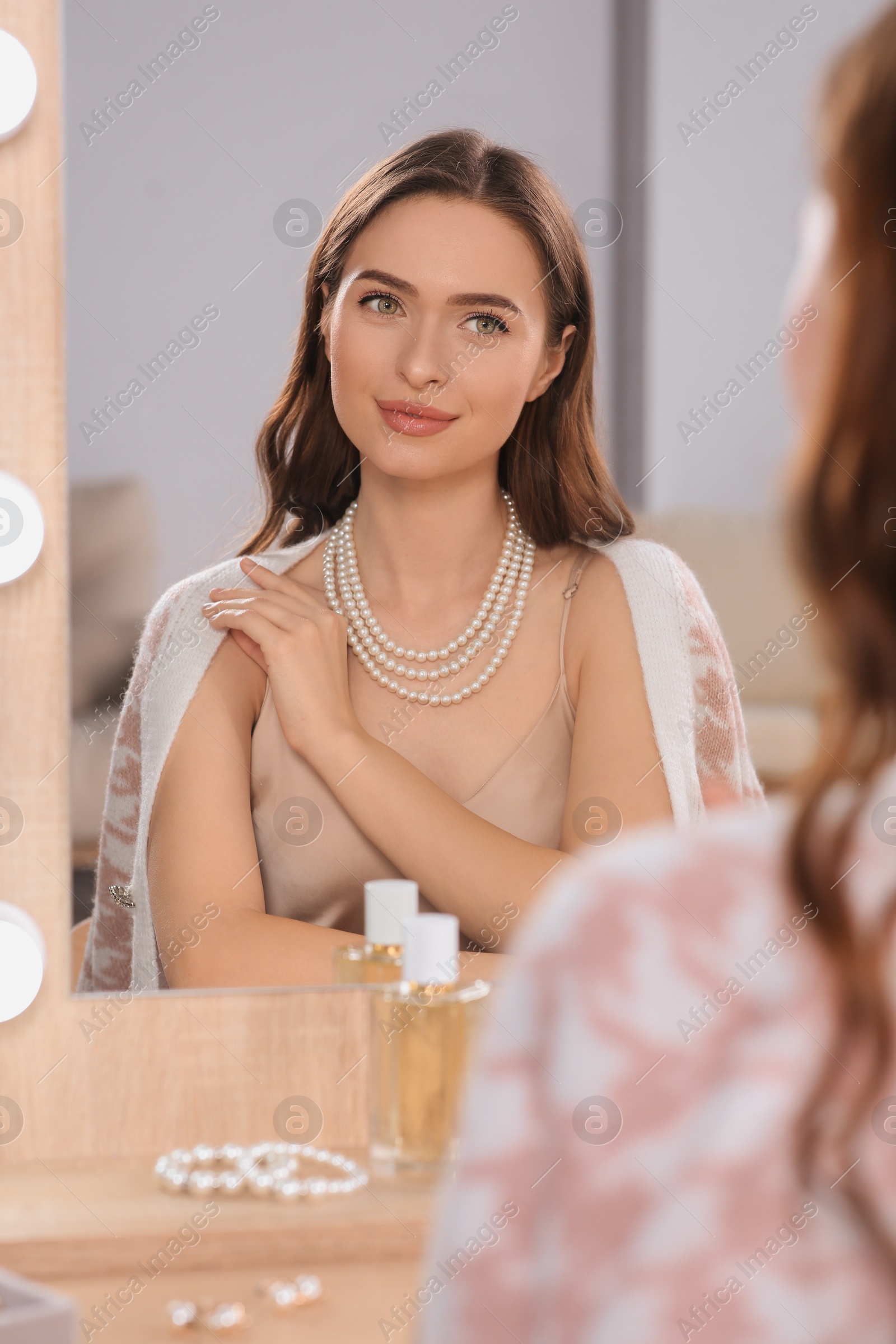 Photo of Young woman wearing elegant pearl necklace near mirror at dressing table indoors