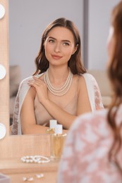Photo of Young woman wearing elegant pearl necklace near mirror at dressing table indoors