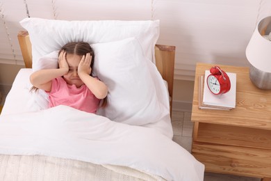 Photo of Little girl covering face in bed and alarm clock on bedside table at home, above view