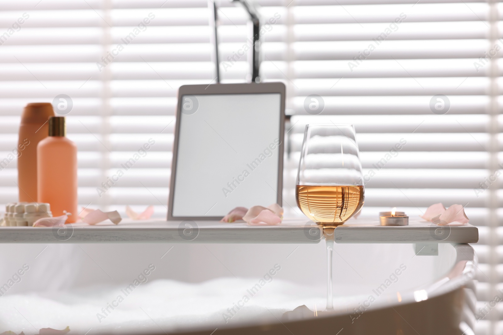 Photo of Wooden tray with tablet, wine, toiletries and flower petals on bathtub in bathroom