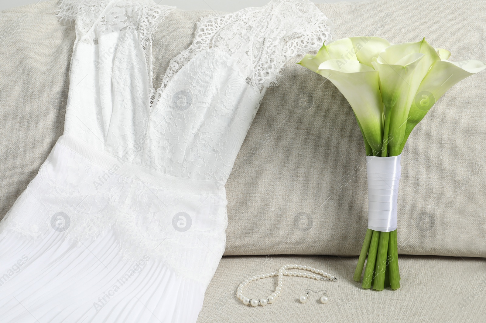 Photo of Beautiful calla lily flowers tied with ribbon, wedding dress and jewelry on sofa