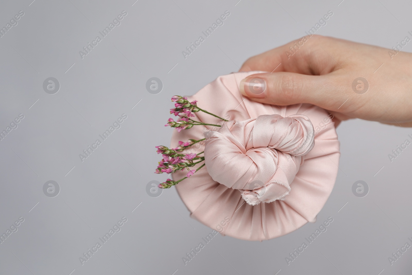 Photo of Furoshiki technique. Woman holding gift packed in fabric and beautiful pink flowers on gray background, closeup with space for text