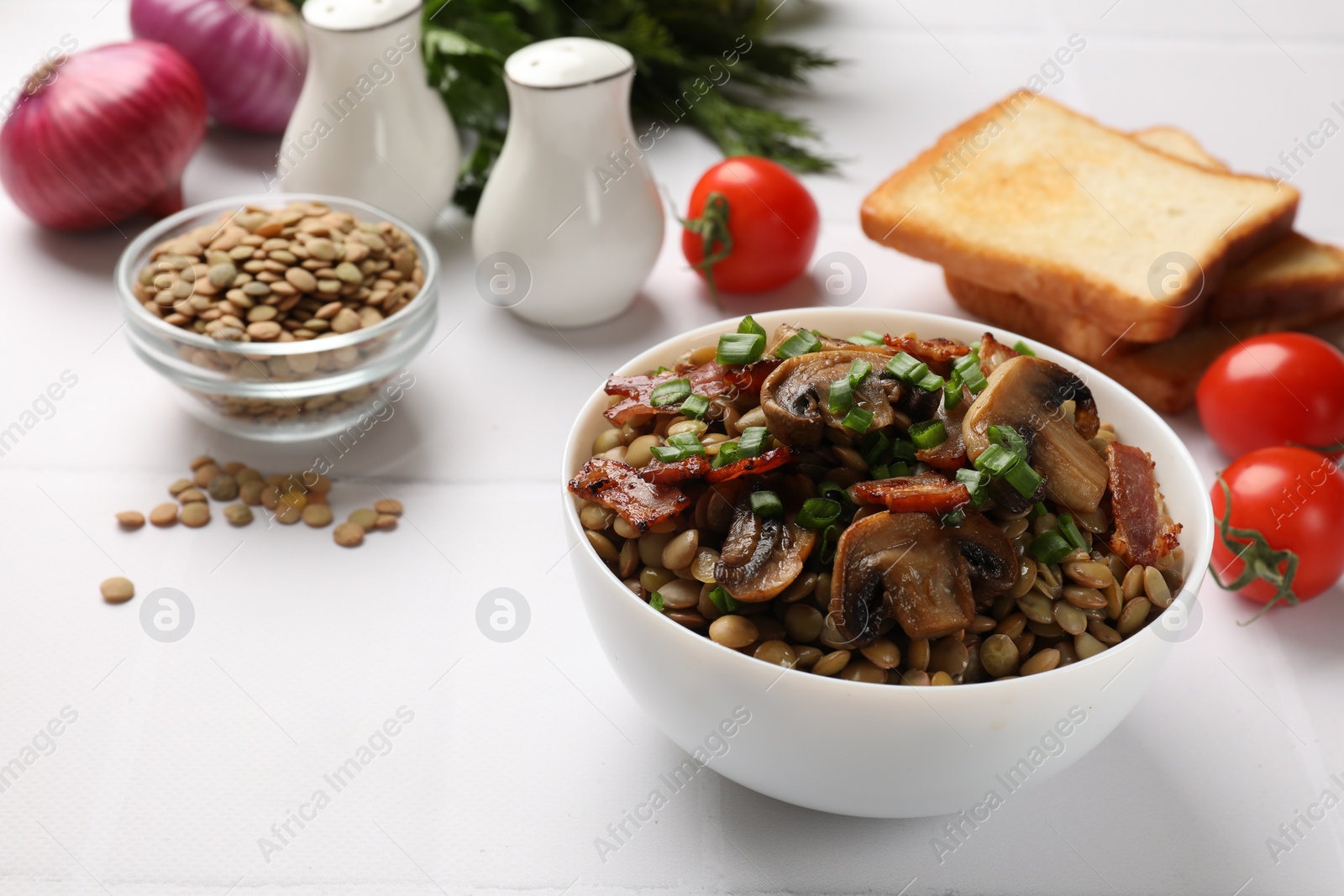 Photo of Delicious lentils with mushrooms, bacon and green onion in bowl served on white table, closeup