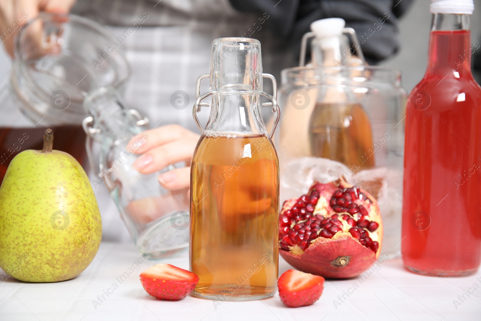 Photo of Making kombucha. Woman pouring drink from jug into bottle at white tiled table, selective focus