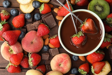 Photo of Fondue forks with strawberries in bowl of melted chocolate surrounded by other fruits on wooden table, flat lay