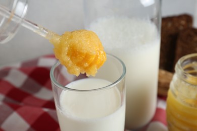 Putting spoon with tasty honey into glass of milk on checkered cloth, closeup