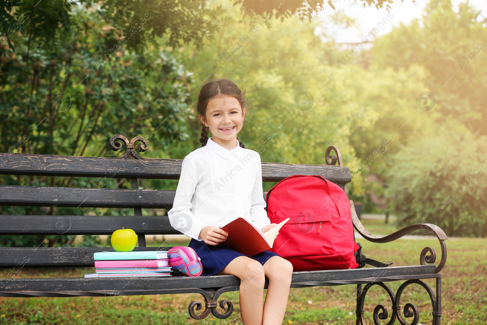 Photo of Cute little school child with stationery reading book on bench in park