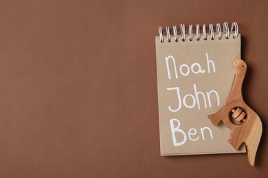 Notebook with list of baby names and wooden toy on brown background, top view. Space for text