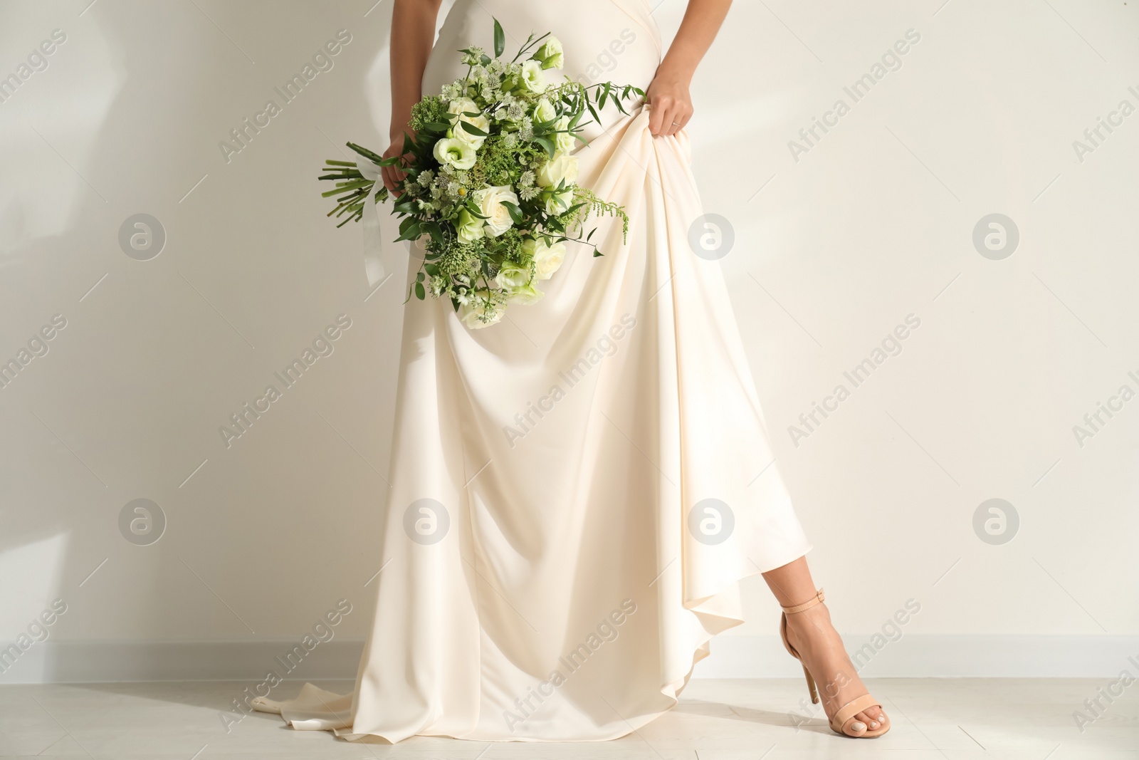 Photo of Young bride wearing wedding dress with beautiful bouquet near light wall, closeup