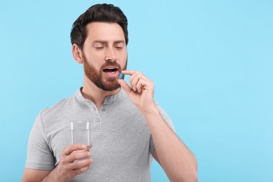 Handsome man with glass of water taking pill on light blue background, space for text