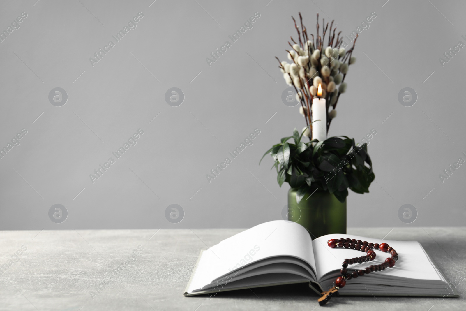 Photo of Burning candle, plant with willow branches, Bible and rosary beads on grey table, space for text