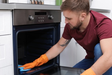 Photo of Young man cleaning oven with rag in kitchen