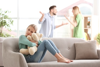 Little unhappy girl sitting on sofa while parents arguing at home