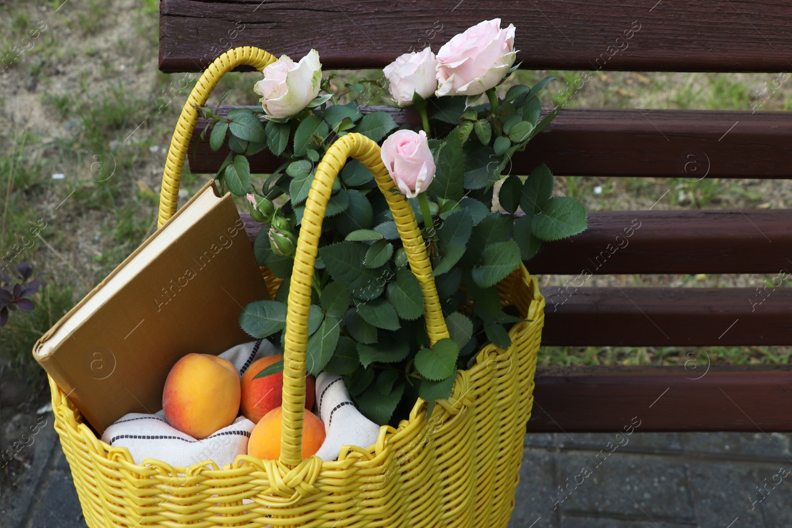 Photo of Yellow wicker bag with roses, book and peaches on bench outdoors