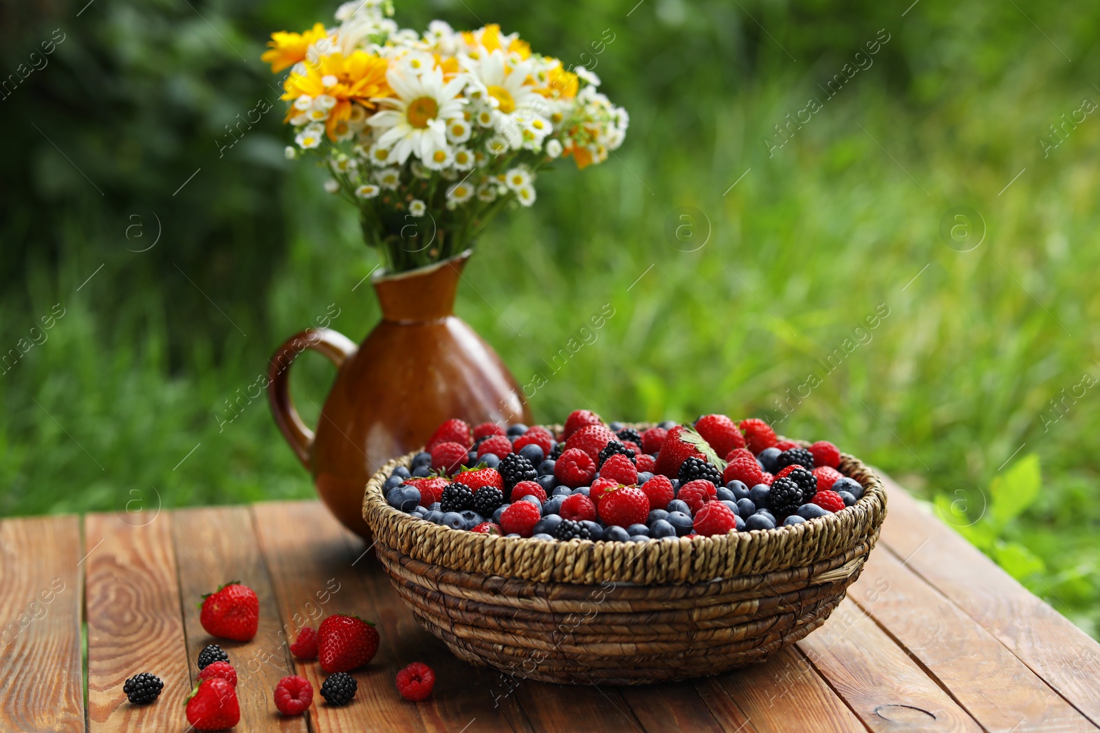 Photo of Wicker bowl with different fresh ripe berries and beautiful flowers on wooden table outdoors