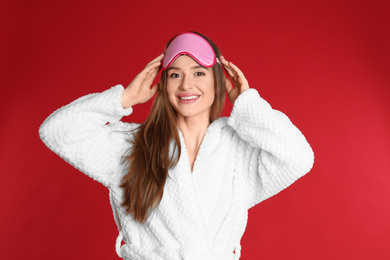 Photo of Young woman in bathrobe and eye sleeping mask on red background