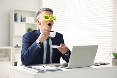 Photo of Man with fake eyes painted on sticky notes holding cup of drink and yawning at workplace in office