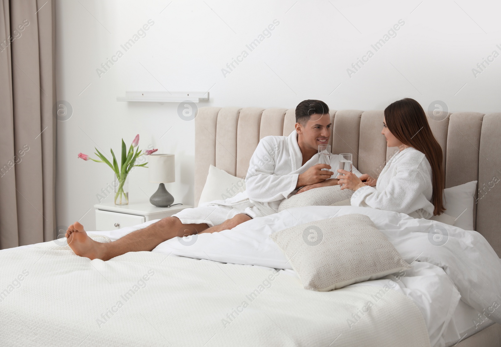 Photo of Happy couple in bathrobes with glasses of water on bed at home