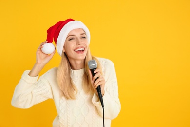 Photo of Emotional woman in Santa Claus hat singing with microphone on yellow background, space for text. Christmas music