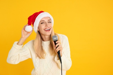 Emotional woman in Santa Claus hat singing with microphone on yellow background, space for text. Christmas music