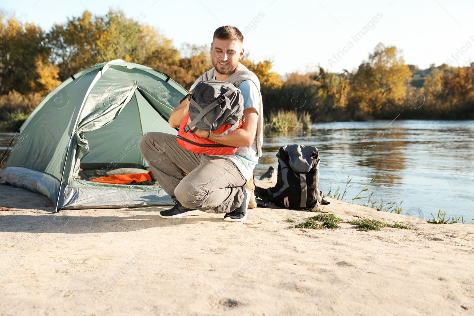 Photo of Young man packing sleeping bag near camping tent outdoors. Space for text