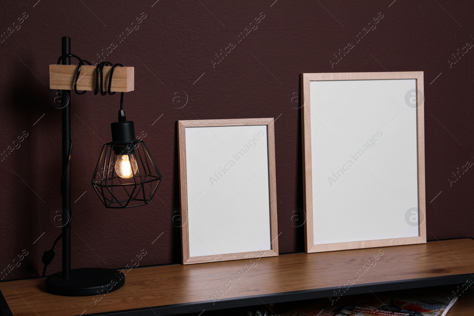 Photo of Empty frames and stylish lamp on wooden table near brown wall. Mockup for design