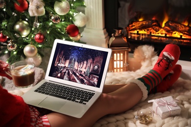 Photo of MYKOLAIV, UKRAINE - DECEMBER 23, 2020: Woman with sweet drink watching Harry Potter and Philosopher's Stone movie on laptop near fireplace at home, closeup. Cozy winter holidays atmosphere