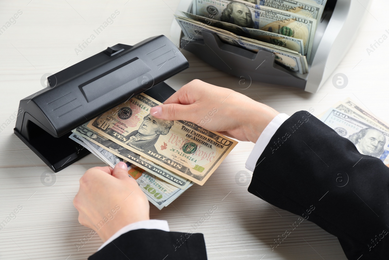 Photo of Woman checking dollar banknotes with currency detector at white wooden table, closeup. Money examination device