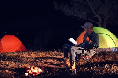Photo of Man with flashlight reading book near bonfire at night. Camping season