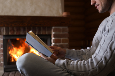 Photo of Man reading book near burning fireplace at home, closeup