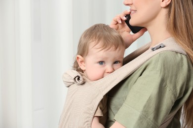 Photo of Mother talking on smartphone while holding her child in sling (baby carrier) indoors