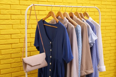 Rack with different stylish clothes and bag near yellow brick wall