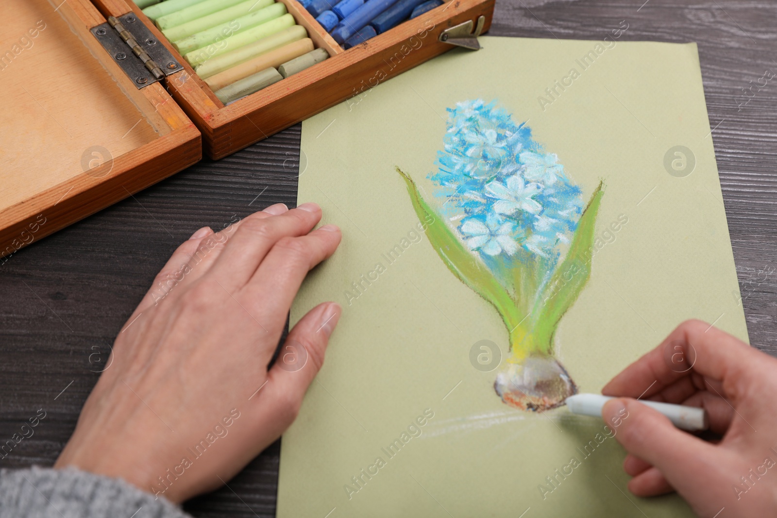 Photo of Woman drawing blue blooming hyacinth on paper with soft pastels at wooden table, closeup